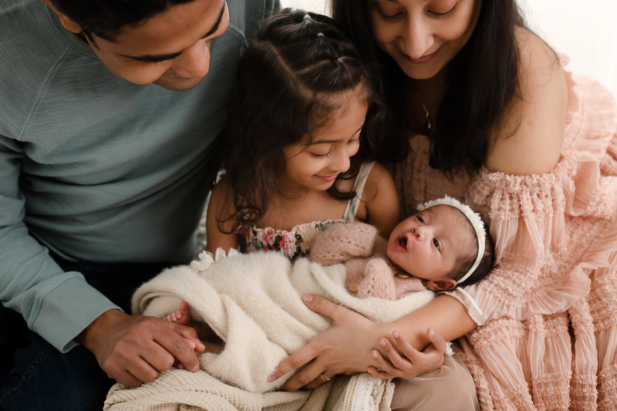 Newborn photographer captures intimate family moment as big sister holds new baby, with proud parents lovingly surrounding their daughters, showcasing the unique storytelling that helps photographers stand out