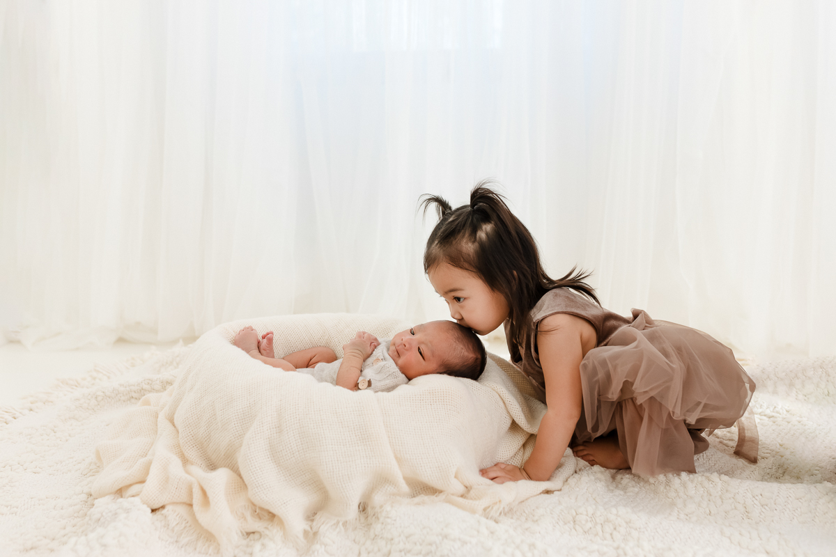 Sweet big sister gently kisses newborn brother's head - the kind of precious moment photographers can capture more often with a repeatable workflow that reduces admin time